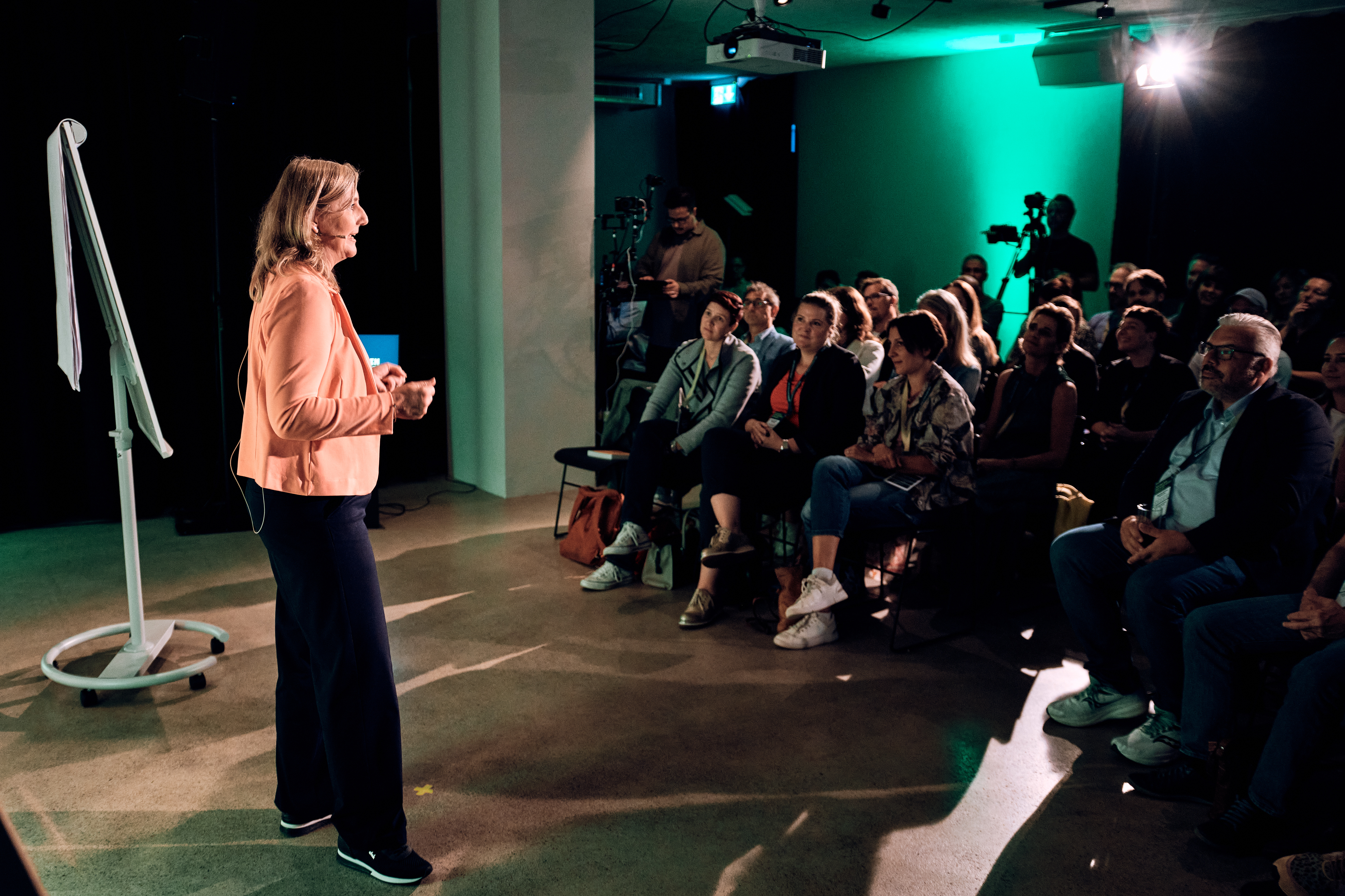 Eine Frau spricht zu einem sitzenden Publikum in einem schummrigen Raum, hinter ihr sind ein Flipchart und eine Filmausrüstung zu sehen.
